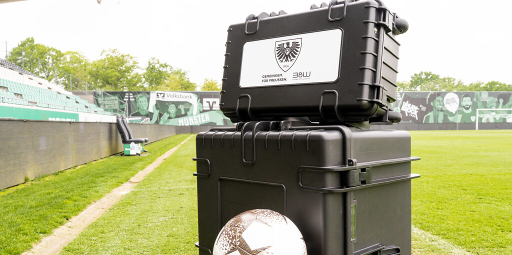 Zwei schwarze B&W International Mehrzweckkoffer mit Vereinslogo des SC Preußen Münster auf einem Fußballfeld.