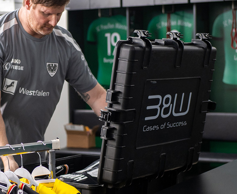 Zeugwart des SC Preußen Münster öffnet schwarzen B&W International Koffer mit Logo im Umkleideraum, bereit für den Einsatz bei Fußballspielen.