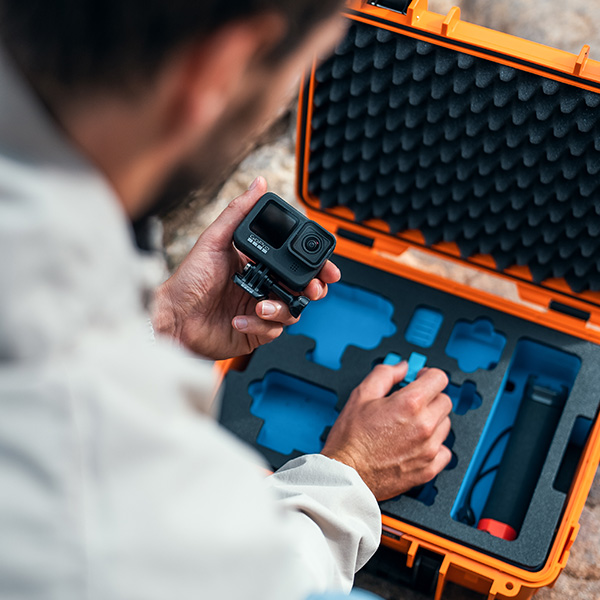 Eine Person hält eine GoPro-Kamera und organisiert das Zubehör in einem orangefarbenen Hartschalenkoffer von B&W International mit Schaumstofffächern.