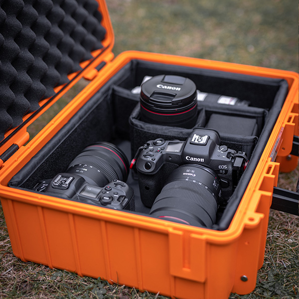 Ein offener orangefarbener Hartschalenkoffer von B&W International, der zwei Canon-Kameras mit angesetzten Objektiven und ein zusätzliches Canon-Objektiv enthält und im Freien auf Gras steht.