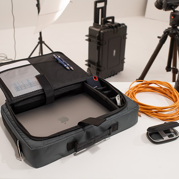 Eine offene Laptoptasche mit einem MacBook, Stiften und Zubehör liegt auf einem Schreibtisch, umgeben von einem B&W International Hartschalenkoffer, einem orangefarbenen Spiralkabel und einer Kameraausrüstung in einer Studioumgebung.