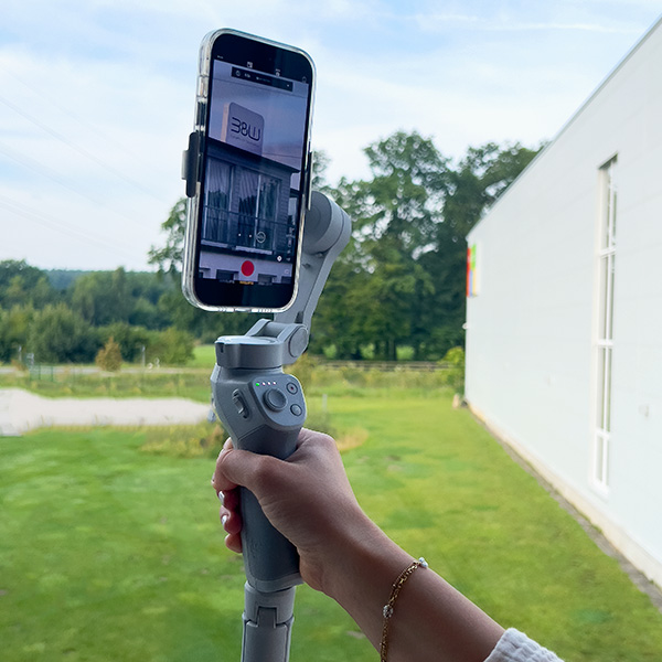 Eine Person hält ein Smartphone, das auf einem Handheld-Gimbal von B&W International montiert ist, und zielt auf ein Gebäude vor einem grasbewachsenen Außenbereich.