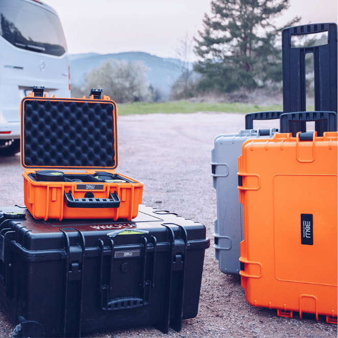 Mehrere B&W International Schutzkoffer in Orange, Schwarz und Grau im Outdoor-Einsatz vor Fahrzeug, ideal für sicheren Transport empfindlicher Ausrüstung.