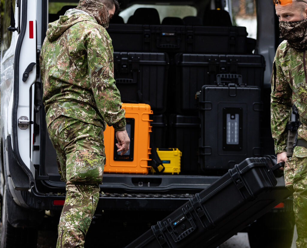 Zwei Personen in Tarnkleidung entladen robuste, schwarz-gelbe Hartschalenkoffer von B&W International aus dem Kofferraum eines Fahrzeugs.