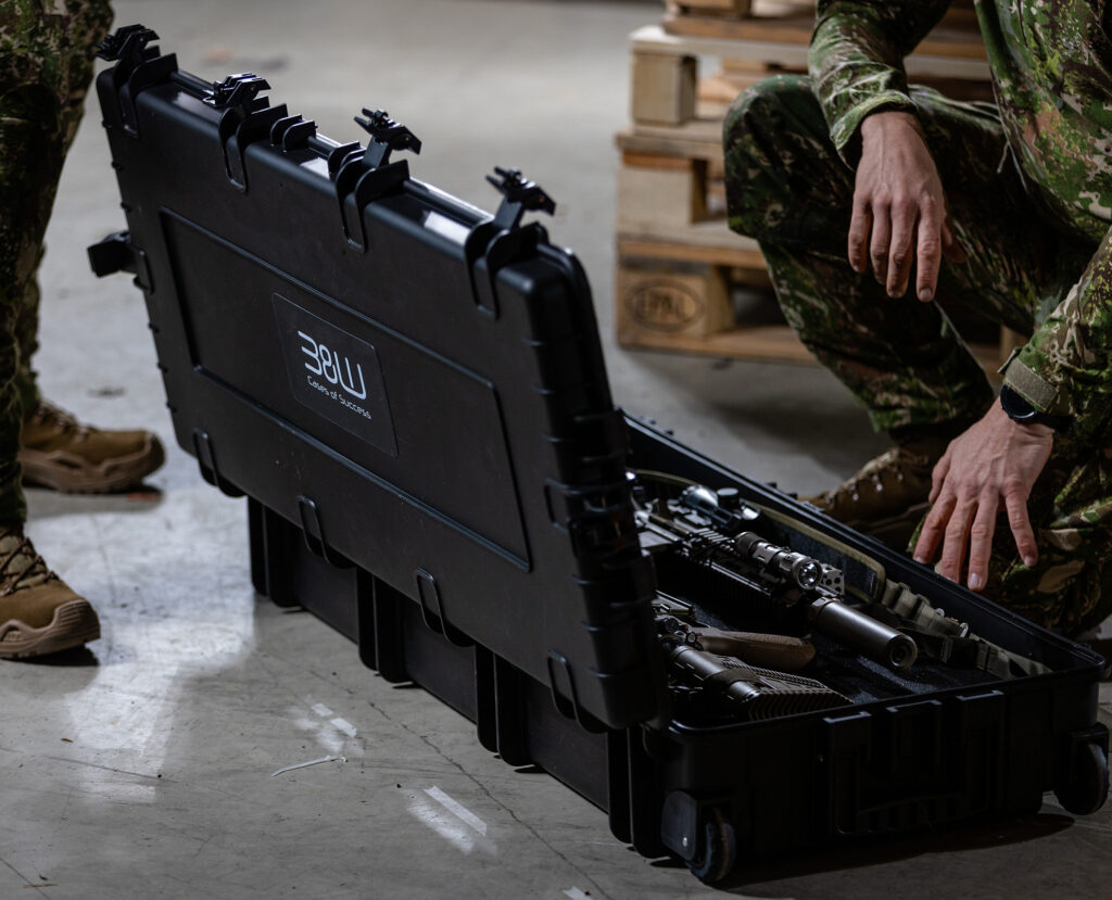Zwei Personen in Tarnhosen knien neben einem geöffneten schwarzen Hartschalenkoffer von B&W International, der Schusswaffen und Ausrüstung auf dem Boden eines Lagerhauses enthält.