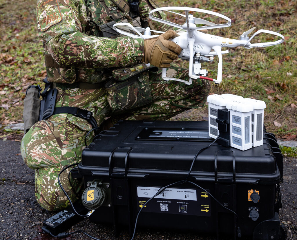 Geschlossener schwarzer Energy Case von B&W International im Vordergrund – im Hintergrund ein Soldat in Camouflage mit weißer Drohne im militärischen Außeneinsatz.