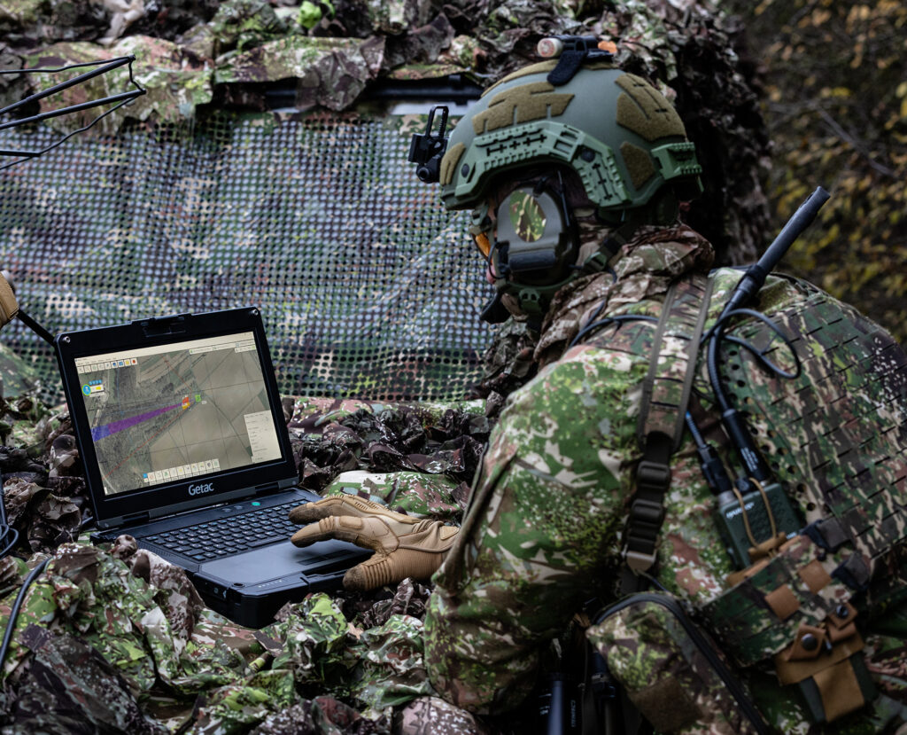 Ein Soldat in Tarnkleidung verwendet einen robusten Laptop von B&W International, der eine Kartenschnittstelle anzeigt, während sich in der Nähe Kommunikationsgeräte in einem Waldgebiet befinden.